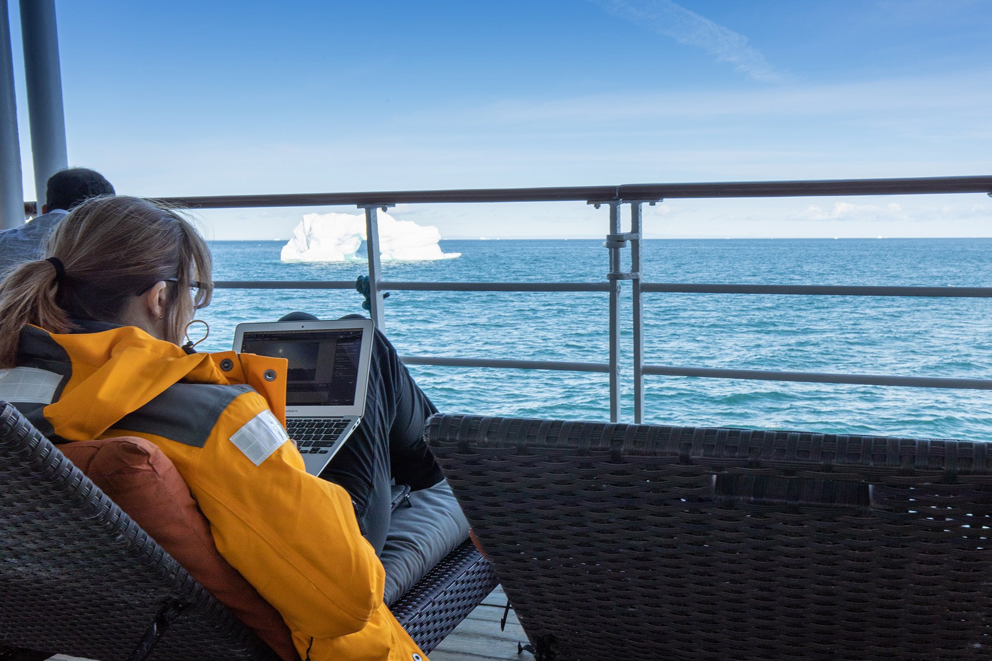 A woman sits on deck of a polar ship with laptop computer while looking out at icebergs 
