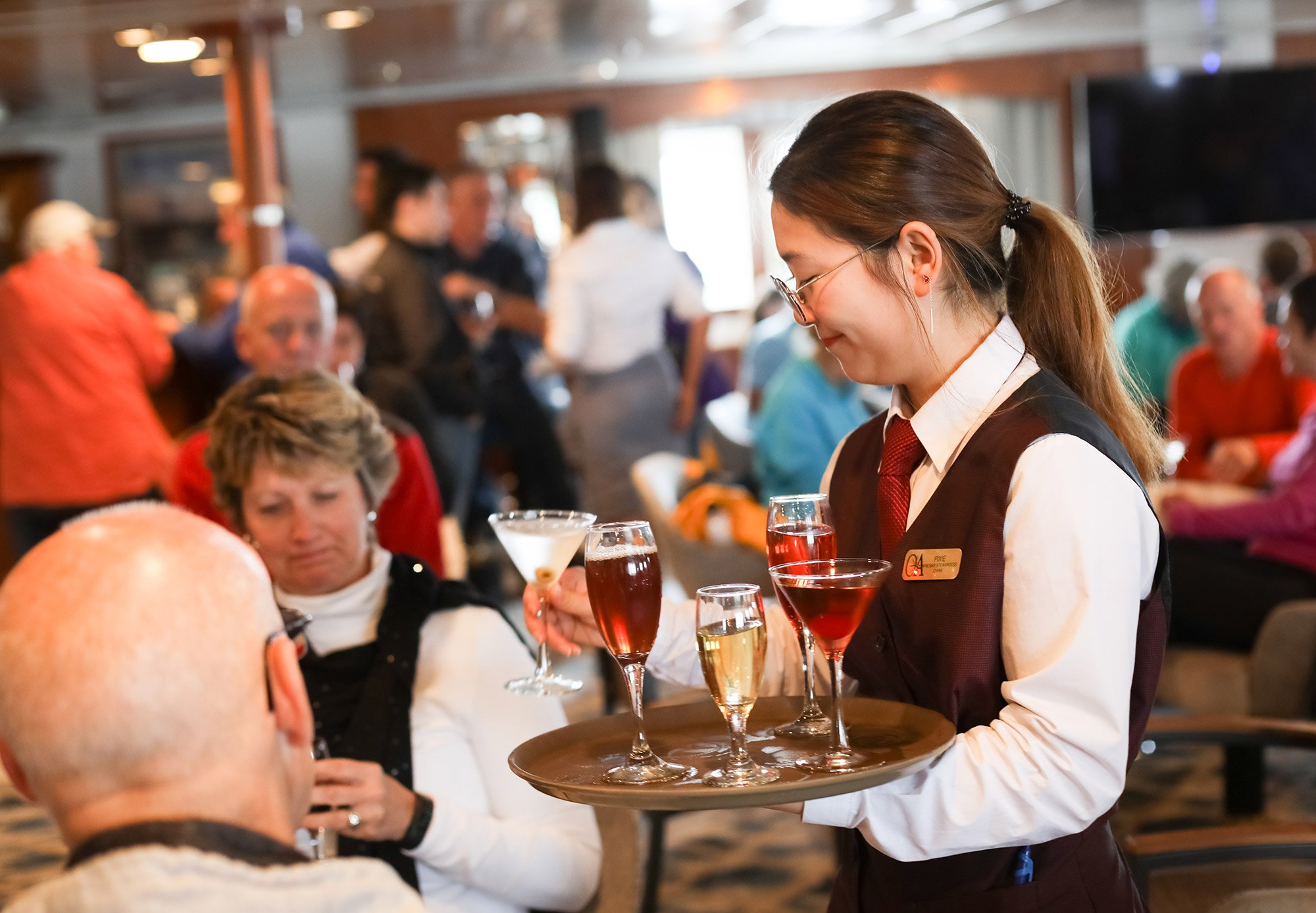 Server with tray of cocktails and alcoholic drinks standing beside seated man and woman on polar expedition cruise ship.