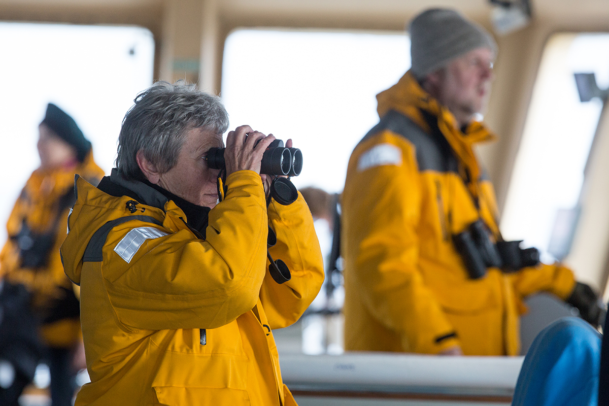 A passenger in a yellow Quark jacket uses binoculars to look out the window on the hull of a ship.