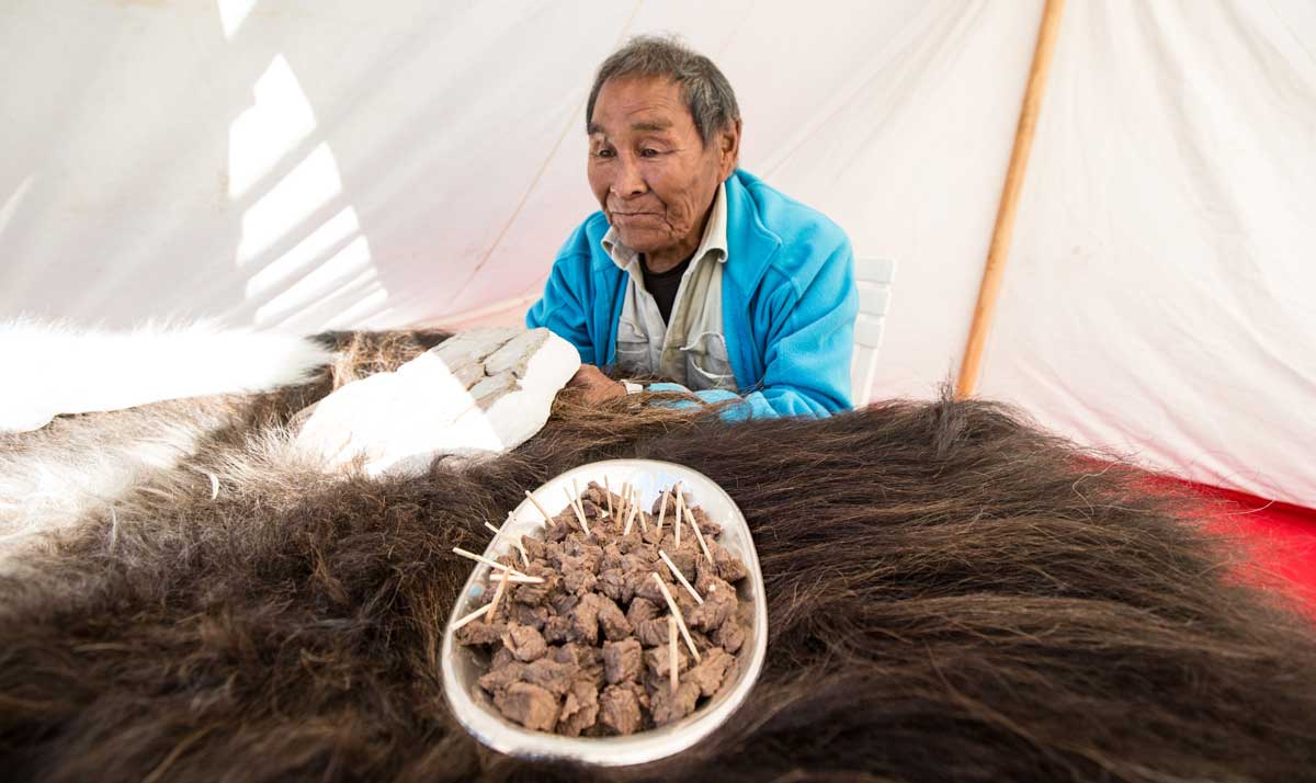 An older man offers meat tidbits as a snack on a bed of furs.