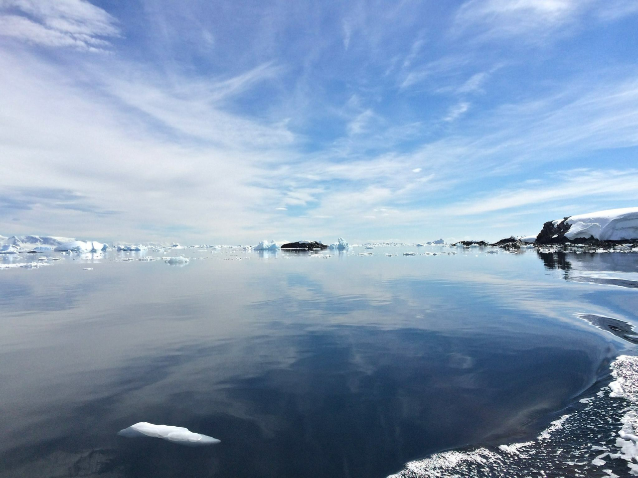 Dreamy blue skies and wispy clouds reflect back over the crystal clear waters of an Antarctic bay, and seen from a cruising Zodiac.
