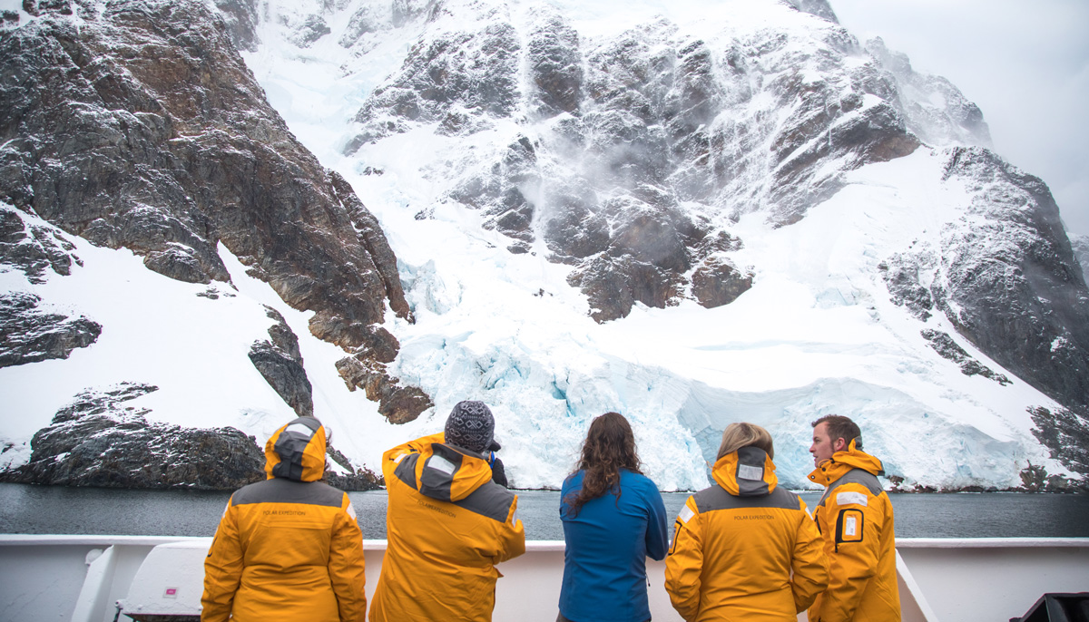 Passengers in yellow jackets stand on a ship and look up at ice covered hills.